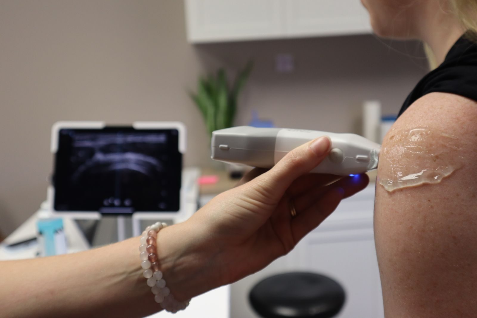 A clinician holding a wireless handheld ultrasound probe against a patient shoulder, with the scan displayed on a tablet in the background